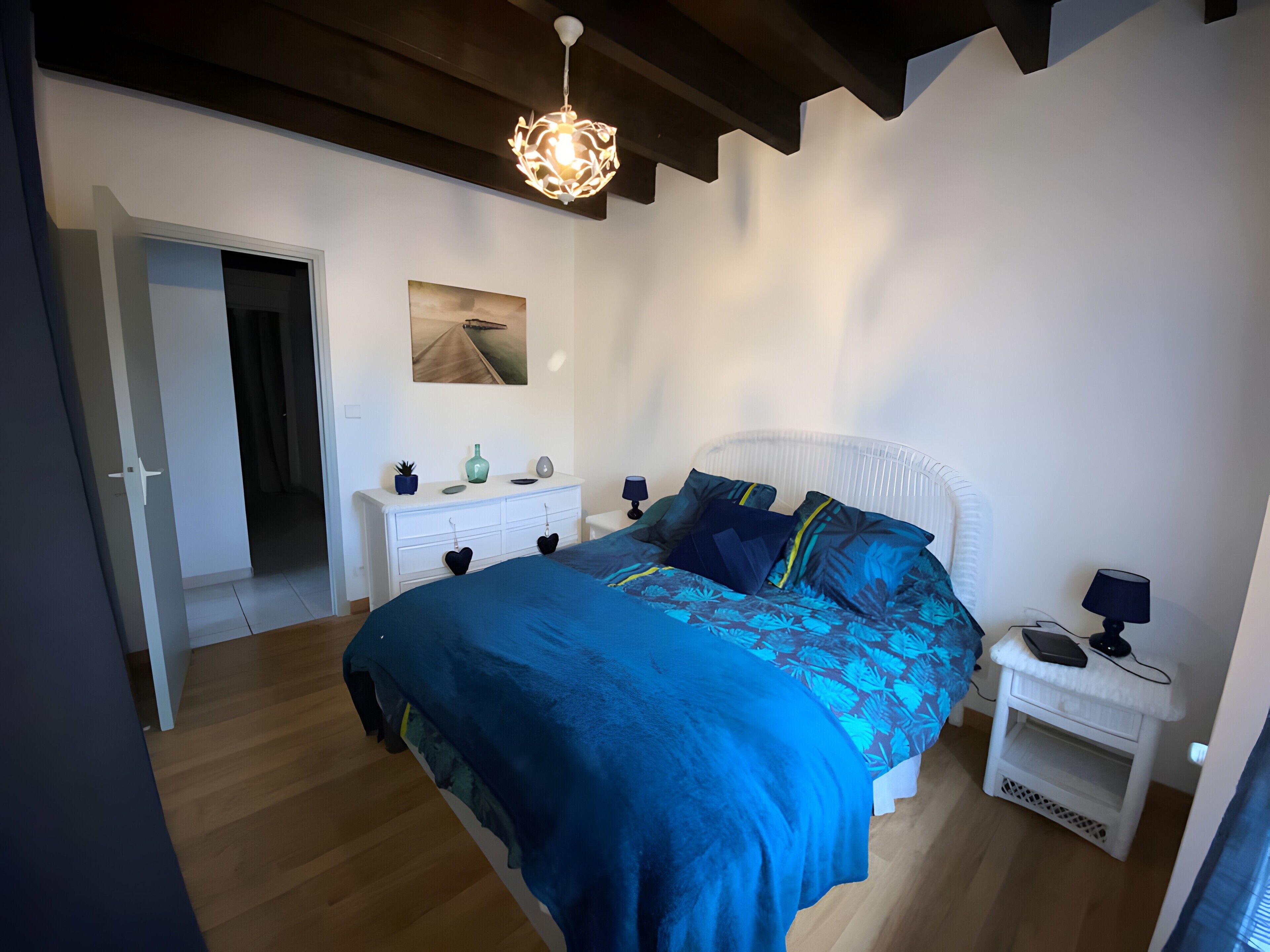 Un dormitorio con cama, mesitas de noche, cómoda, lámpara colgante y un cuadro en la pared.