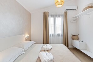 A hotel room with a bed, a bedside table, a hanging light fixture, a window with curtains, and an air conditioning unit. - Estay - Aura Mare -  The charm of the sea in Arbatax (Arbatax)