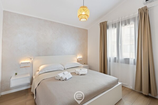 A modern hotel room with a large bed, bedside lamps, a small table with a plant, and a window with curtains. - Estay - Aura Mare -  The charm of the sea in Arbatax (Arbatax)