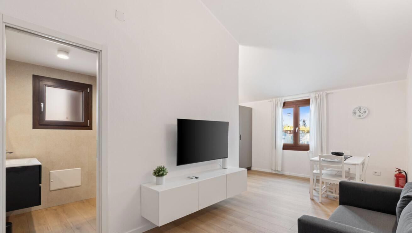 A modern living room with a flat-screen TV on a white console, a dining area with a table and chairs, and a view of the outside through a window.