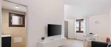 A modern living room with a flat-screen TV on a white console, a dining area with a table and chairs, and a view of the outside through a window.