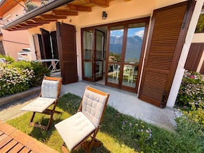 A modern outdoor seating area with two cushioned chairs, a wooden deck, and a view of mountains through large glass doors. - La Quiete 28: Swimming Pool and View (Tremosine)