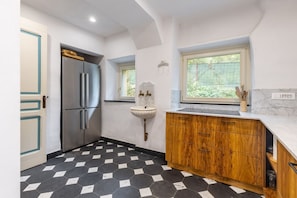 A kitchen with a black and white hexagonal tile floor, a stainless steel refrigerator, wooden cabinets, and a window with a view of greenery. - Hostdomus - Villa Celestia (Alassio)