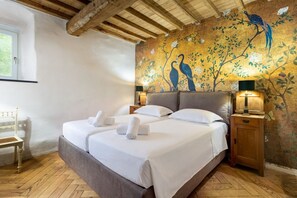 Un llit doble amb llençols blancs, una tauleta de nit de fusta amb una làmpada, i un mural de pássars a la paret.