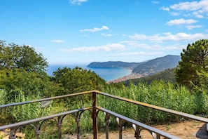 A scenic view from a balcony with a railing, overlooking a coastal landscape with trees and a distant town. - Hostdomus - Villa Celestia (Alassio)