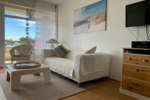 Ein Zimmer mit einem weißen Sofa, einem kleinen weißen Tisch, einem Korbstuhl, einem Holzschrank und einem Fernseher.