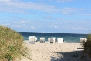 Ein Strand mit weißen Liegestühlen, die am Sand aufgereiht sind, einem klaren Himmel und dem Meer im Hintergrund.