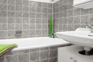 A bathroom with a bathtub, a sink, and grey tiled walls. - “Beach Break” at Haus Hamburg (Großenbrode)