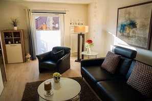 Ein Wohnzimmer mit einem schwarzen Ledersofa, einem weißen Couchtisch, einem kleinen Beistelltisch mit Lampe, einer Topfpflanze und einem Gemälde an der Wand.