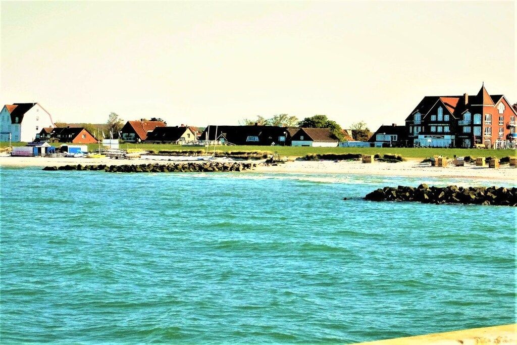 Un paysage côtier avec des maisons situées près de l’eau, une jetée et un grand bâtiment doté d’un toit rouge.
