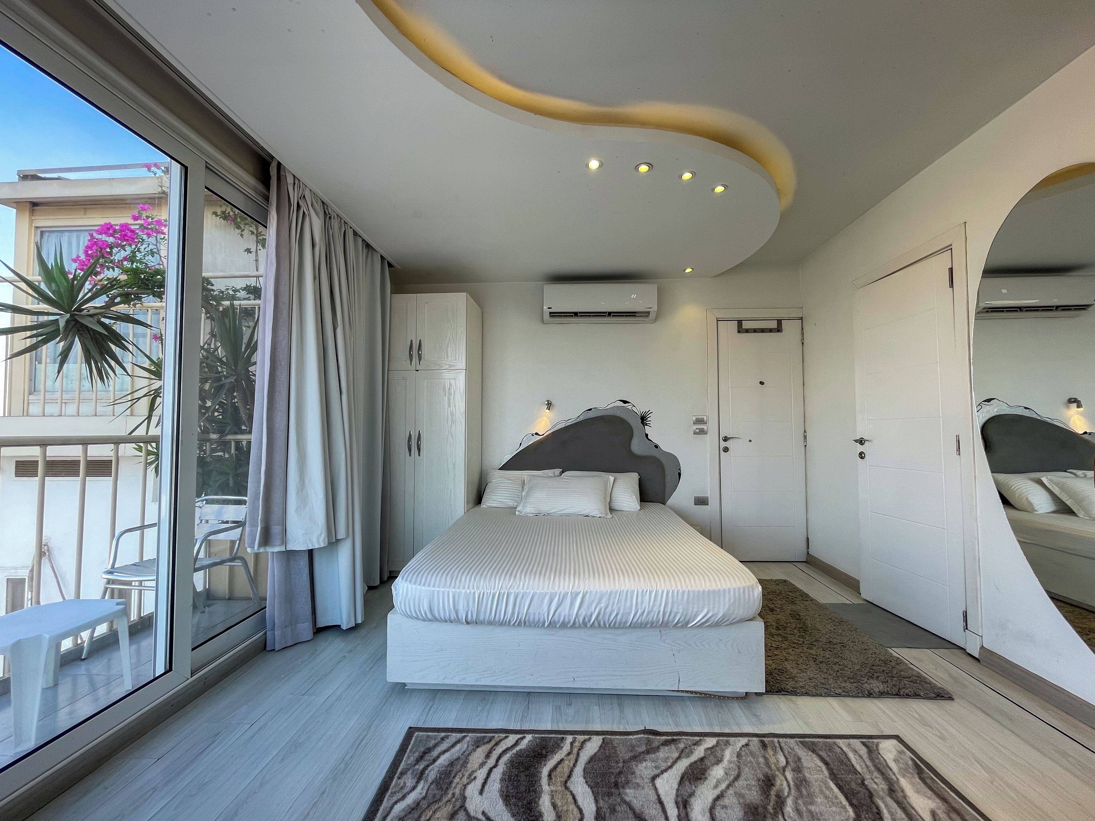 A modern bedroom with a large bed, a sliding glass door to a balcony, and a unique ceiling design.