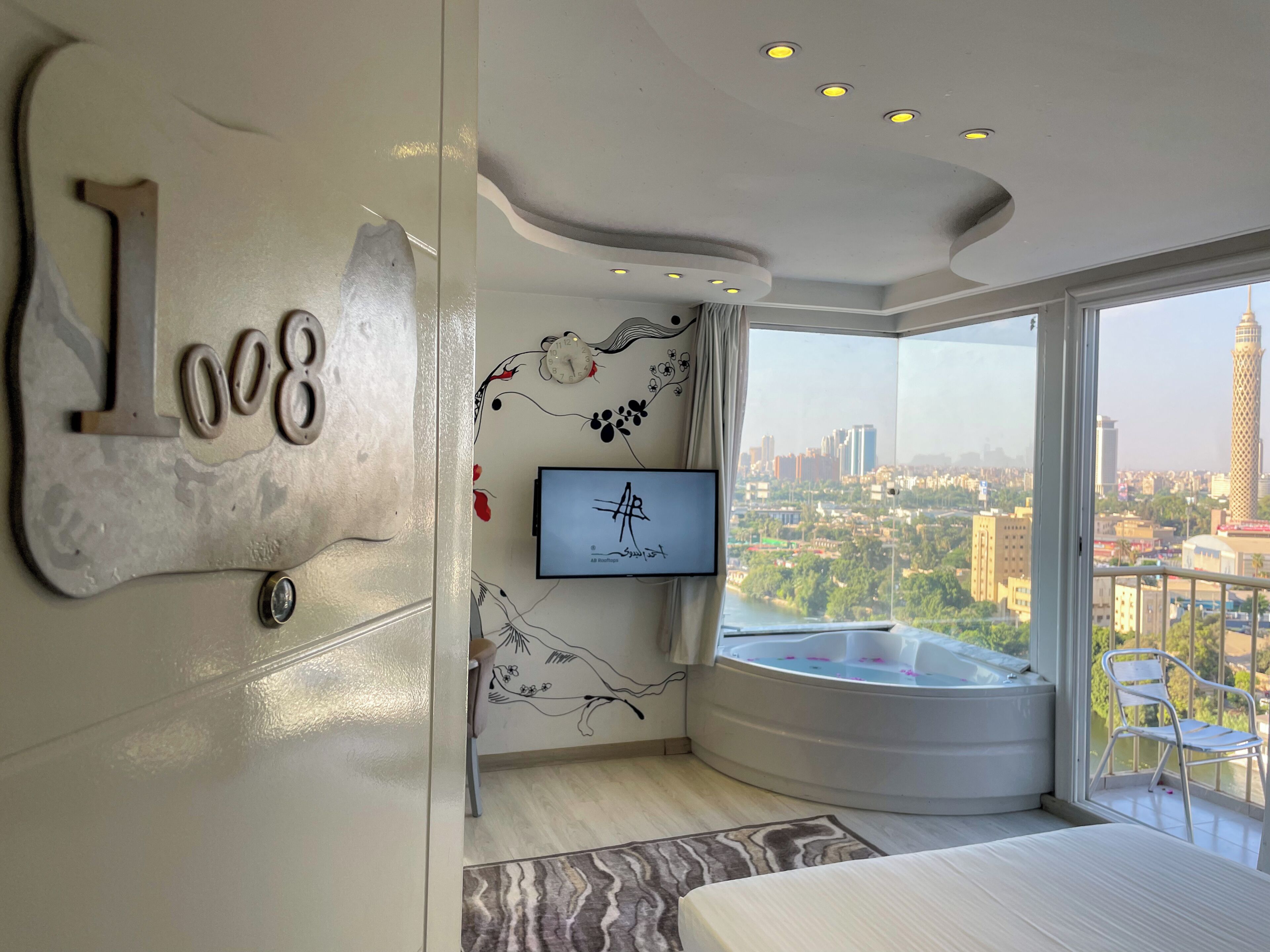 Habitació d'hotel moderna amb un llit gran, una televisió penjada a la paret, una placa decorativa amb un número, un balcó amb vista a la ciutat i una banyera d'hidromassatge.