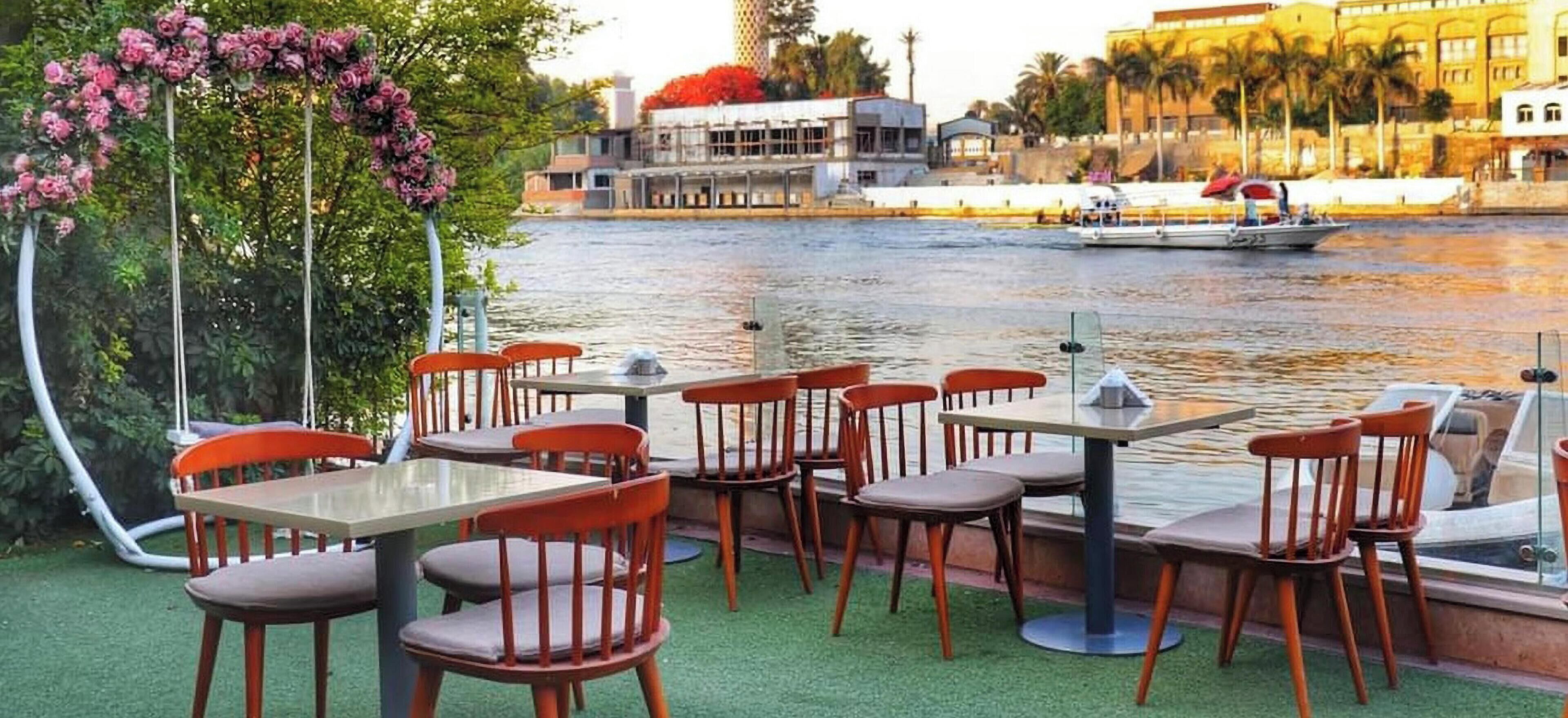 An outdoor seating area with tables and chairs facing a river, with a building in the background.