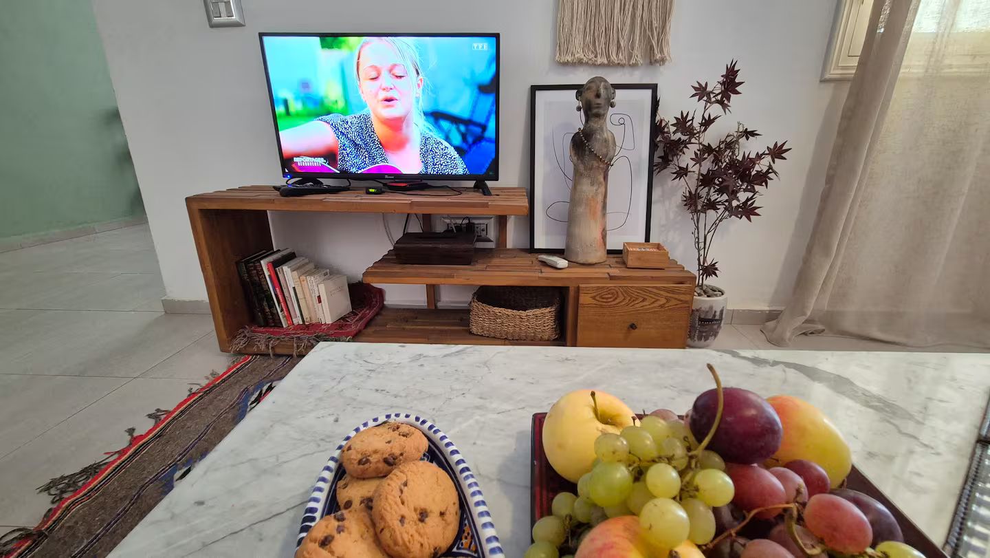 A living room with a television on a wooden stand, a vase with decorative branches, and a table with cookies and fruit.
