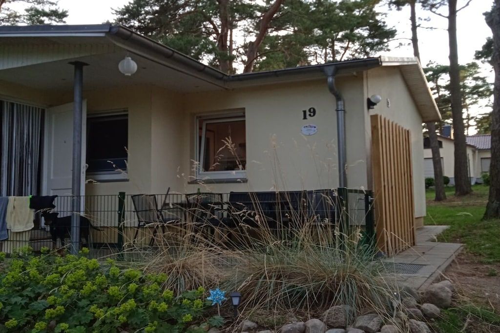 A single-story building with a number 19 sign, a porch with a chair, and a small garden area with rocks and plants.