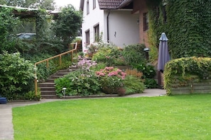 Ein Garten mit Wohnbebauung, eine Treppe führt zu einem Haus, umgeben von üppigem Grün und blühenden Pflanzen.