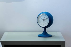 A blue clock with a white face and black numbers on a white table against a plain wall. - Pop Urban Escape near the center (Athens)