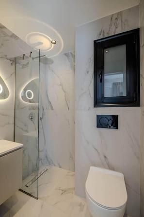 A modern bathroom with a glass shower enclosure, a white toilet, and a marble wall. - Tiny colorful studio near the Historic Center (Athens)