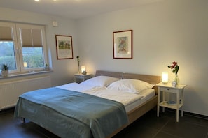 Une chambre à coucher avec un lit, des tables de chevet, une fenêtre avec des stores et des tableaux encadrés accrochés au mur.