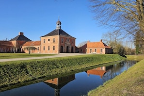 Un grand bâtiment en briques, doté d’une tour centrale, qui se reflète dans une étendue d’eau calme, sous un ciel dégagé.