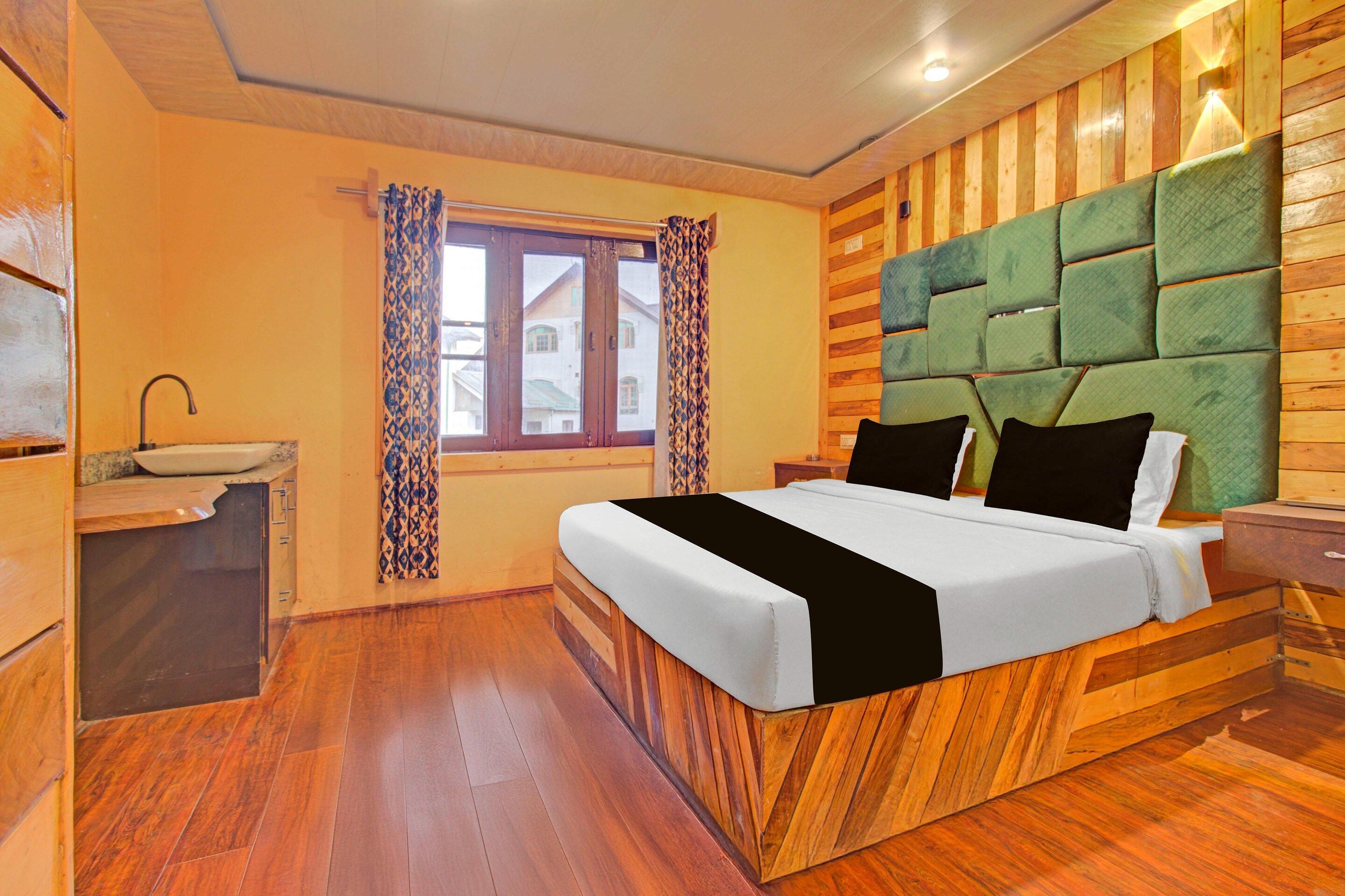 A hotel room with a wooden floor, a bed with a green upholstered headboard, a bedside table, and a window with patterned curtains.
