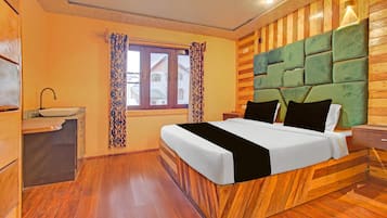 A hotel room with a wooden floor, a bed with a green upholstered headboard, a bedside table, and a window with patterned curtains.