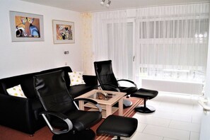 A modern living room with a black leather sofa, two black armchairs, a glass coffee table, and a small black ottoman. There are two framed artworks on the wall, and the room has a large window with sheer curtains. - Holiday apartment with 1 bedroom (Schönberger Strand)