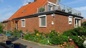 Uma casa de dois andares de tijolo, com telhado de telhas vermelhas, uma varanda com guarda-corpo metálico e um jardim com diversas flores.