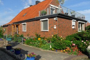 A two-story brick house with a red-tiled roof, a balcony with a metal railing, and a garden with various flowers. - Holiday apartment with 1 bedroom (Ratzeburg)