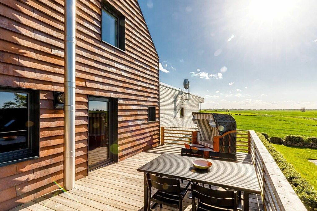 Une maison moderne avec une façade en bois, une terrasse dotée de mobilier d’extérieur et une vue sur un champ verdoyant.