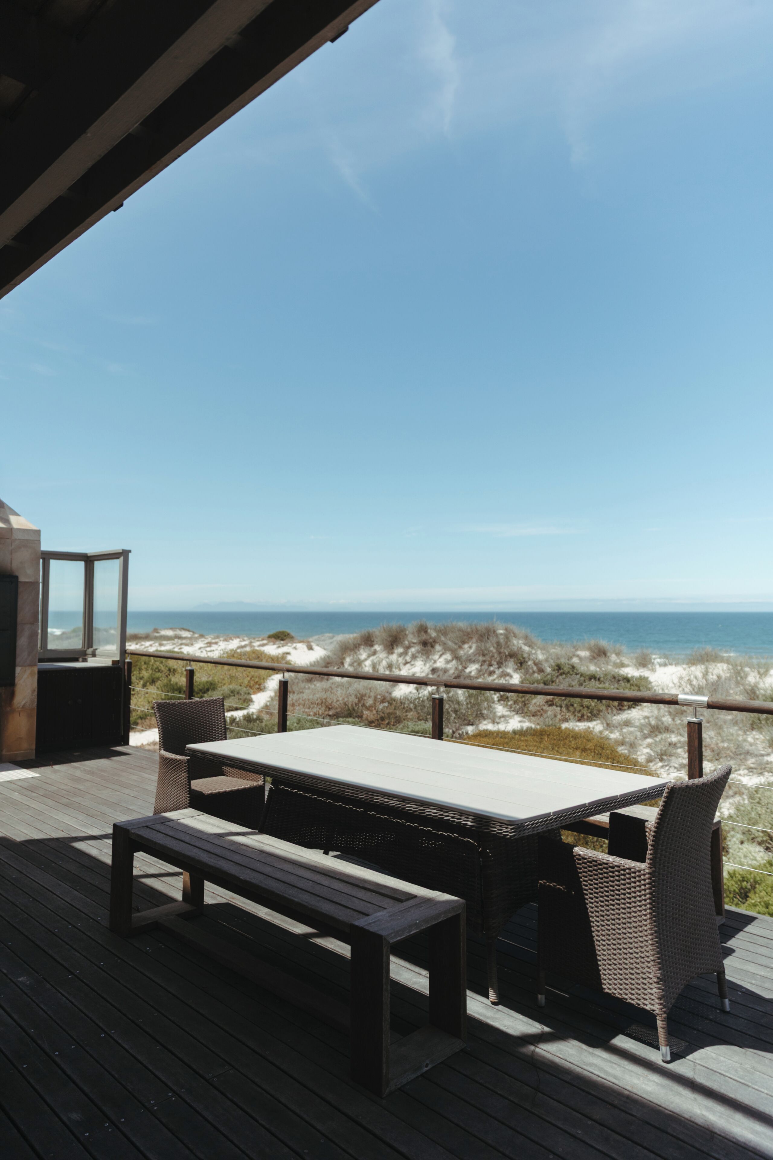 An outdoor deck with a table and chairs, overlooking a beach with dunes and the ocean.
