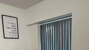 Un comedor con una mesa y sillas blancas, un atril de metal negro con plantas en macetas, una ventana con persianas y una cita inspiradora en la pared.