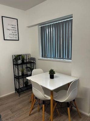 Un coin repas avec une table et des chaises blanches, un présentoir à plantes en métal noir avec des plantes en pot, une fenêtre avec des stores et une citation inspirante sur le mur.