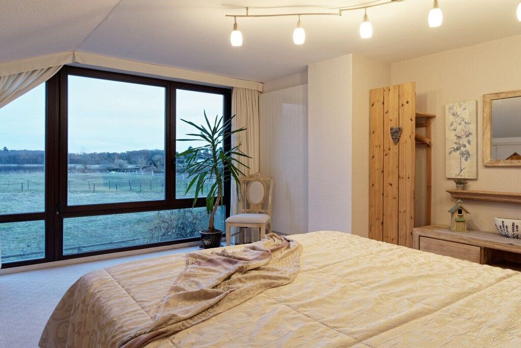 A bedroom with a large window, a bed, a chair, a wooden wardrobe, and a small table with a decorative item.