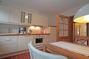 A compact kitchen with white cabinets, a stove, and a dining table with a striped tablecloth. - Holiday apartment with 2 bedrooms (Krummhörn-Greetsiel)