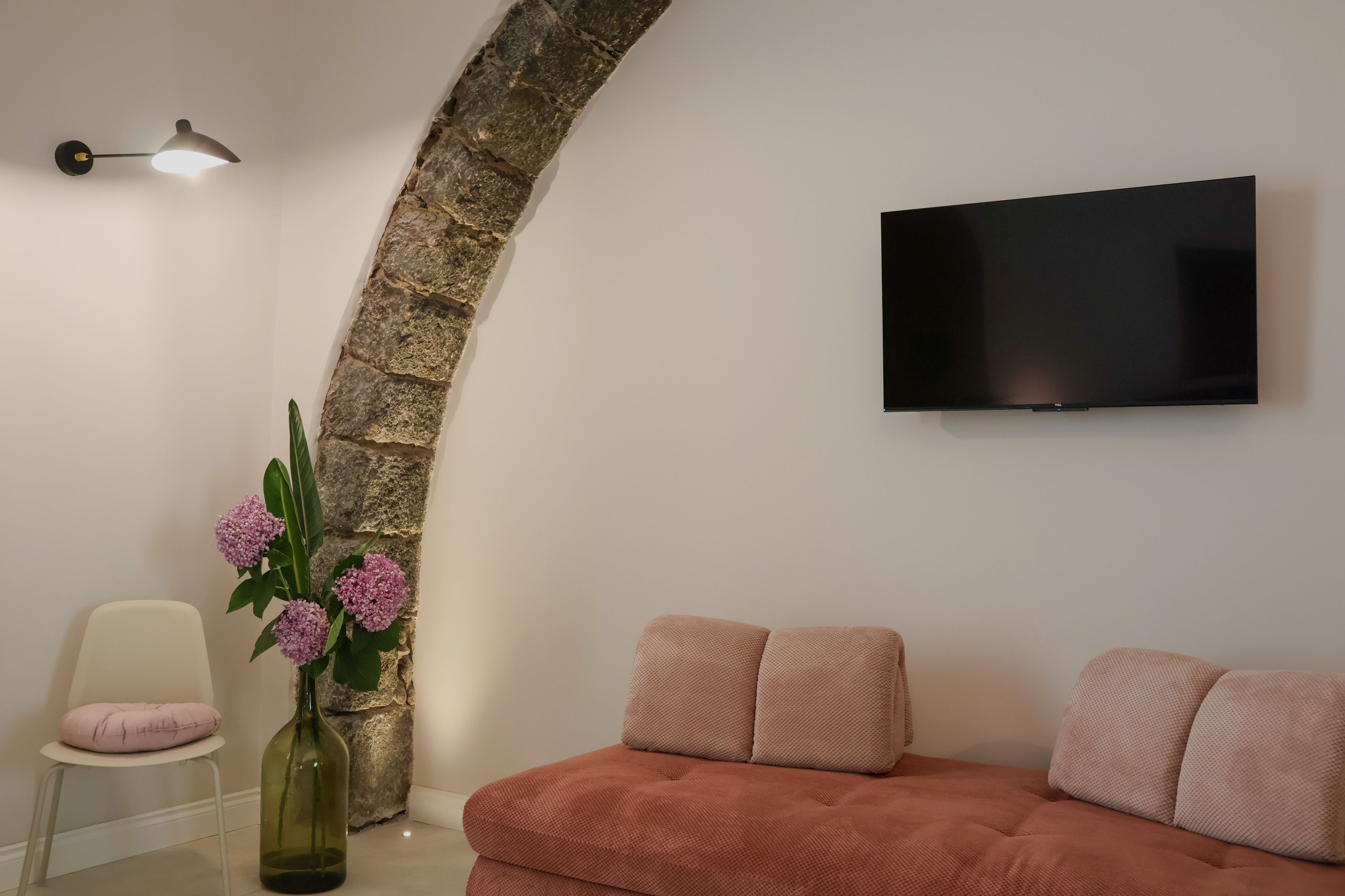 Un soggiorno moderno con un arco in pietra, un televisore a schermo piatto montato a parete, un divano rosa, una poltrona bianca e un vaso con fiori.