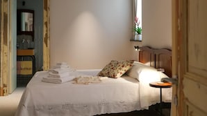 Un dormitorio con cama, mesita de noche, ventana con planta y puerta que da a otra habitación.