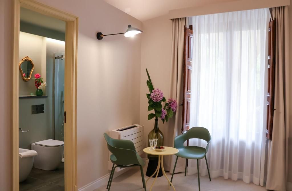 Una habitación con baño, una pequeña mesa con sillas, un florero con flores y una ventana con cortinas.