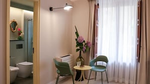 Una habitación con baño, una pequeña mesa con sillas, un florero con flores y una ventana con cortinas.
