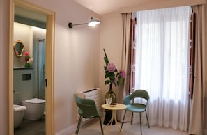 A room with a bathroom, a small table with chairs, a vase with flowers, and a window with curtains. - LA BARONESSA (Riposto)