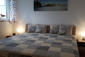 Une chambre à coucher avec un lit, une table de chevet, une lampe, un tableau accroché au mur et une fenêtre avec des rideaux.