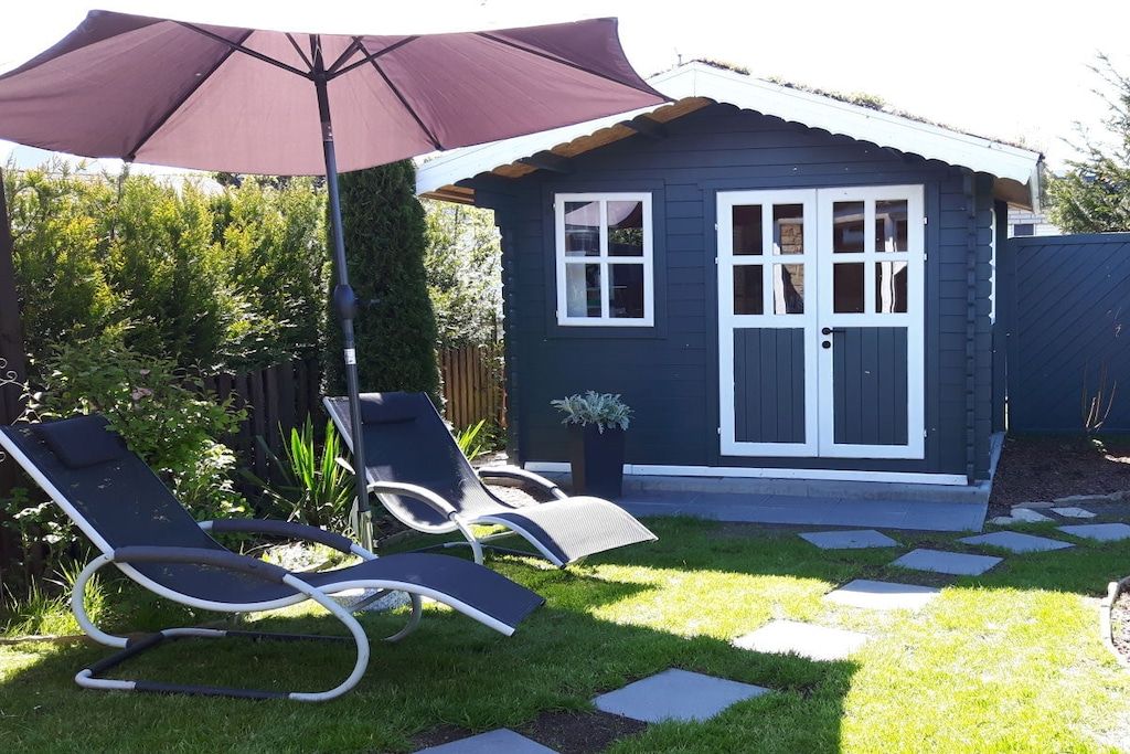 A small house with a dark exterior, a white door, and a window. There are two lounge chairs and a large umbrella in the yard.