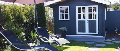 A small house with a dark exterior, a white door, and a window. There are two lounge chairs and a large umbrella in the yard.