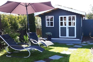A small house with a dark exterior, a white door, and a window. There are two lounge chairs and a large umbrella in the yard. - Holiday home with 2 bedrooms (Butjadingen)