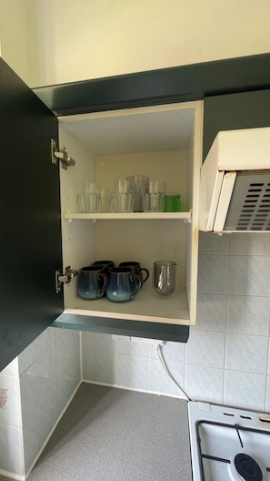 A kitchen with a white tiled backsplash, a black countertop, and a white cabinet with shelves holding clear glasses and blue mugs.