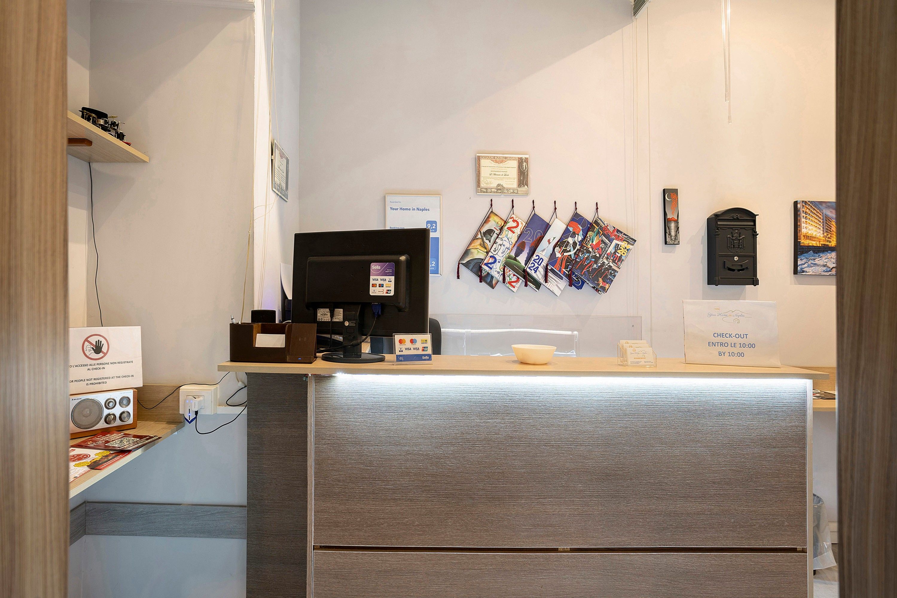 A hotel reception desk with a computer, a check-out counter, and various informational posters on the wall.
