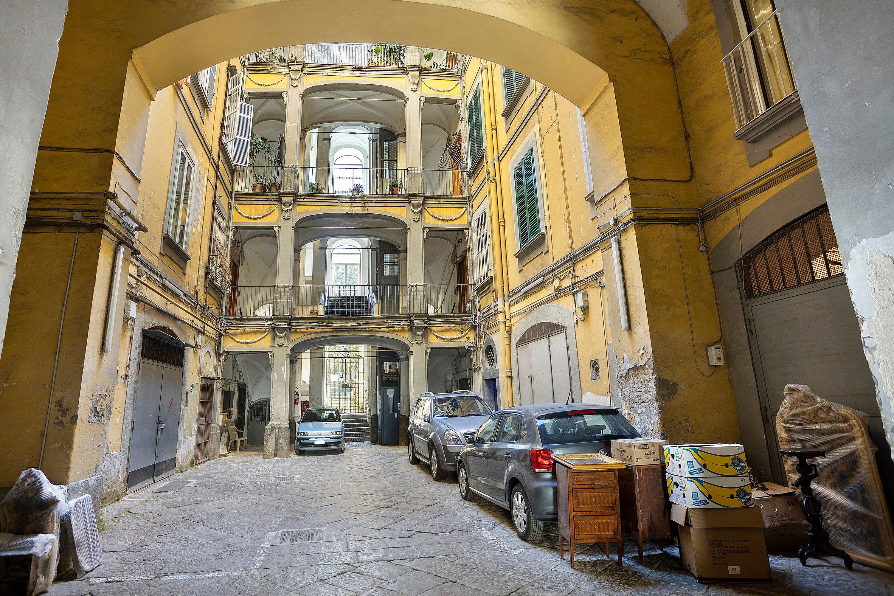 A narrow street with parked cars and construction materials, leading to a building with multiple balconies and a central archway.