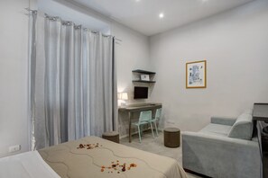 A modern bedroom with a bed, a desk, two chairs, a small round ottoman, and a framed picture on the wall. - Deluxe Room with Balcony, Wi-Fi & Air Conditioning (Napoli)