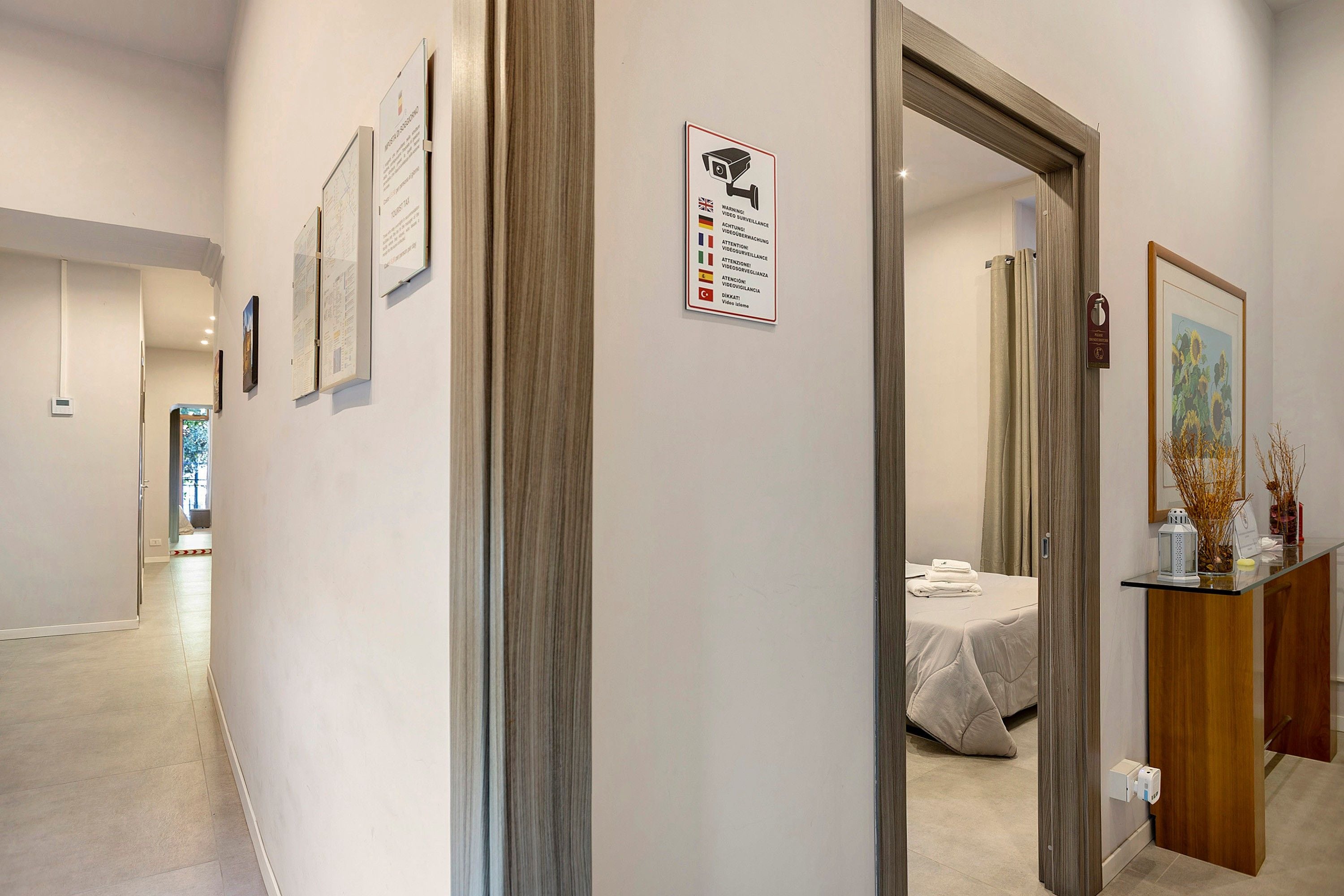 A hotel corridor with framed informational posters, a security warning sign, and a glimpse of a bedroom with a bed and a nightstand.