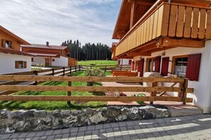 A wooden balcony with a stone wall in the foreground, a grassy area with a bench, and a view of a forest in the background. - Holiday home in Allgäu with 3 bedrooms (Lechbruck am See)
